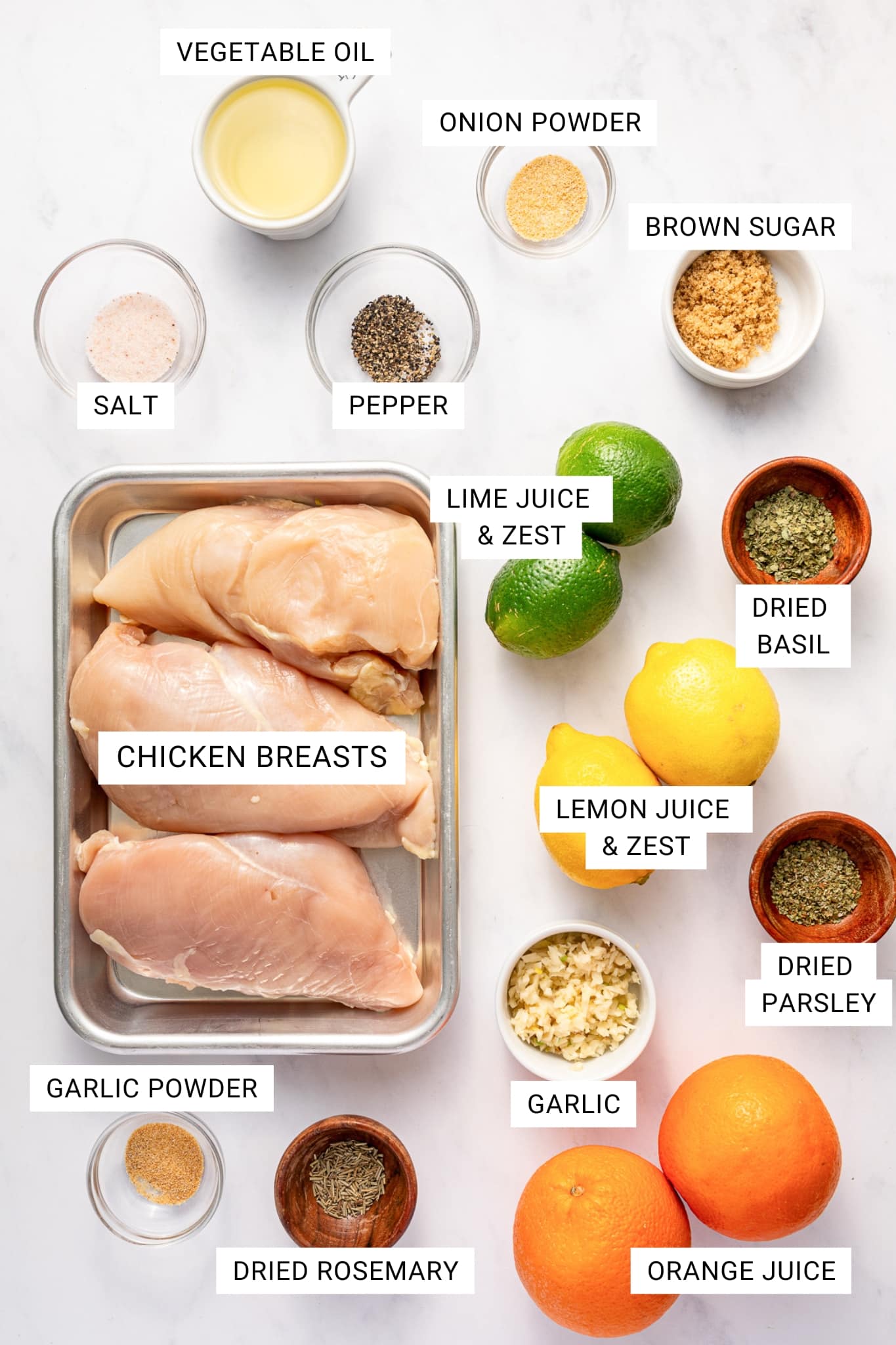 Top-down ingredient shot for citrus chicken marinade on a white marble background with labeled text boxes over each ingredient. A metal tray in the center holds three raw boneless skinless chicken breasts. Surrounding it are whole limes labeled lime juice and zest, whole lemons labeled lemon juice and zest, whole oranges labeled orange juice, a white ramekin of minced garlic labeled garlic, a white measuring cup of pale yellow vegetable oil labeled vegetable oil, small glass bowls of salt, pepper, garlic powder, and onion powder, a white bowl of brown sugar, and small wooden bowls of dried basil, dried parsley, and dried rosemary. No garnish is added beyond the whole citrus fruits.