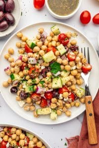 Close-up of Mediterranean Chickpea Salad on a white plate with canned chickpeas, diced cucumber, halved cherry tomatoes, red onion, Kalamata olives, feta cheese, parsley, and black pepper, with a fork, small bowl of dressing, tomatoes, olives, and a rust-colored napkin around the plate.