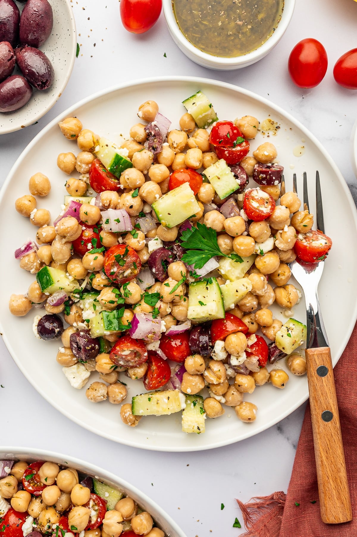 Close-up of Mediterranean Chickpea Salad on a white plate with canned chickpeas, diced cucumber, halved cherry tomatoes, red onion, Kalamata olives, feta cheese, parsley, and black pepper, with a fork, small bowl of dressing, tomatoes, olives, and a rust-colored napkin around the plate.