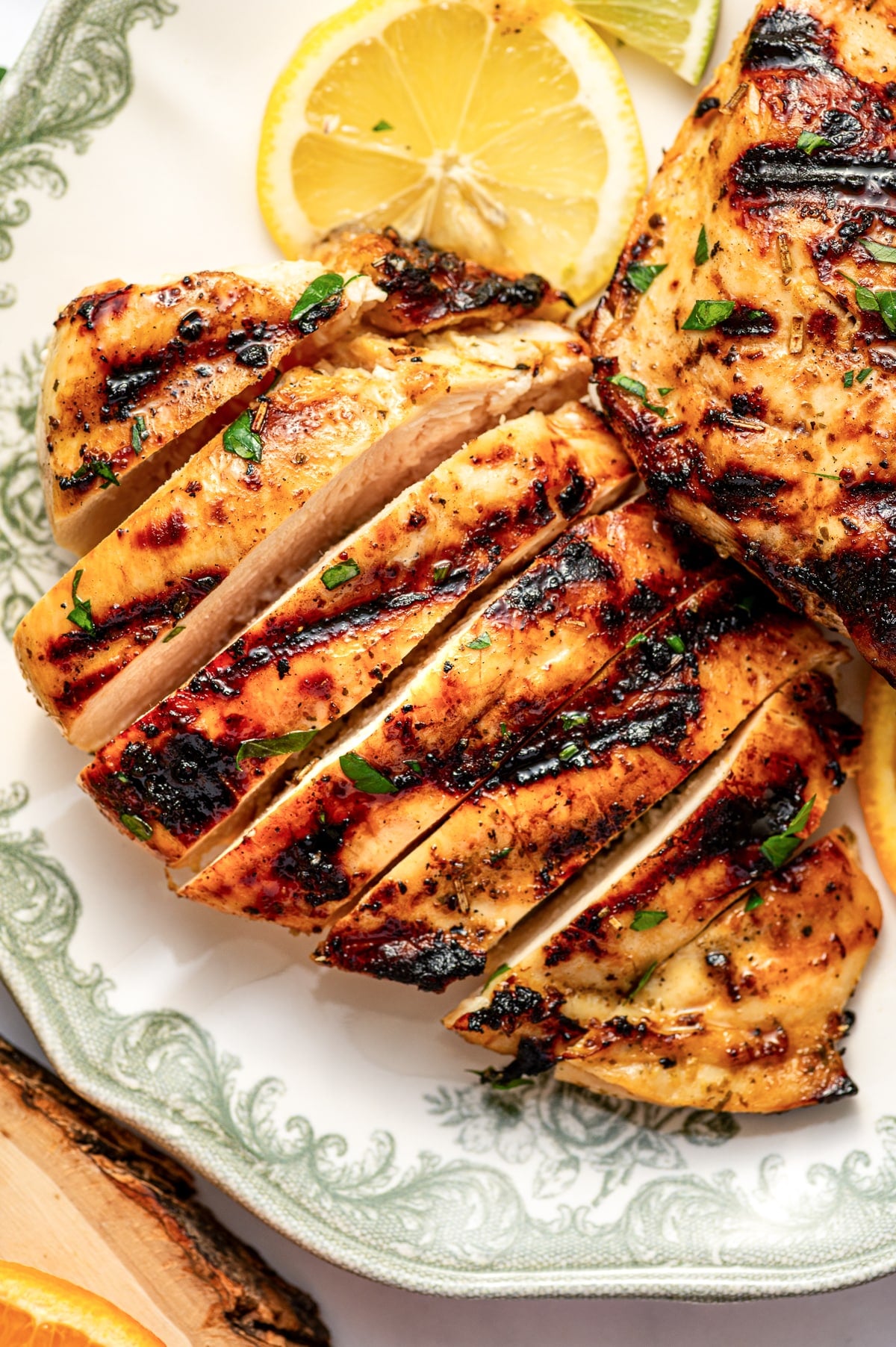 Close-up overhead view of sliced grilled citrus marinated chicken breast on a plate with a sage green decorative border. The chicken is cut into thick slices and shows a juicy interior with dark charred grill marks on the outside. Chopped parsley is scattered over the chicken, and part of a lemon slice and another grilled chicken breast are visible in the background. A rustic wood slice peeks in at the lower left edge.