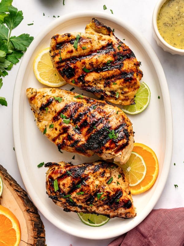 Top-down view of three grilled citrus marinated chicken breasts on a large oval off-white platter. The chicken has dark grill marks and is garnished with chopped green parsley. Lemon, lime, and orange slices are tucked around the chicken. A small off-white ramekin of pale yellow citrus marinade sits in the upper right corner. Fresh parsley, citrus halves, a rustic wood slice, and a dusty rose pink linen napkin surround the platter on a white background.