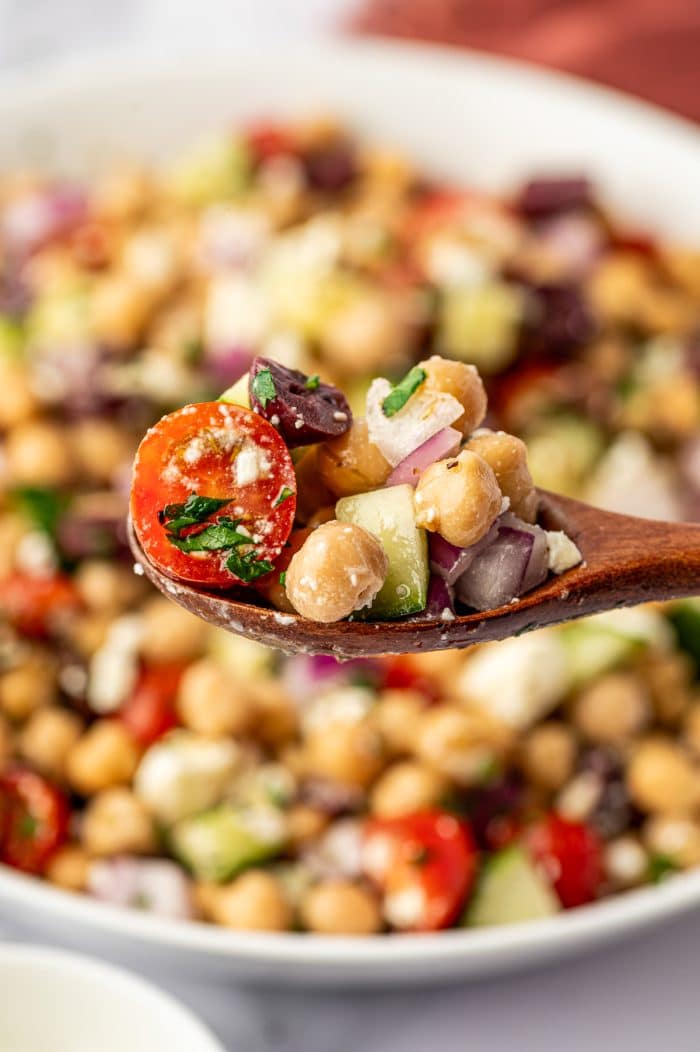 Close-up of Mediterranean Chickpea Salad on a dark wooden spoon with chickpeas, cucumber, cherry tomato, red onion, Kalamata olive, parsley, and crumbled feta cheese, with the salad blurred in the background.
