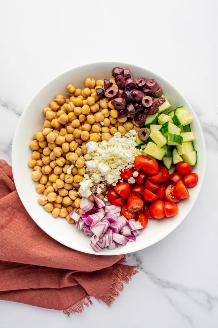 Mediterranean Chickpea Salad ingredients arranged in a large white bowl with canned chickpeas, diced cucumber, halved cherry tomatoes, chopped red onion, sliced Kalamata olives, and crumbled feta cheese on a white marble surface with a rust-colored linen napkin.