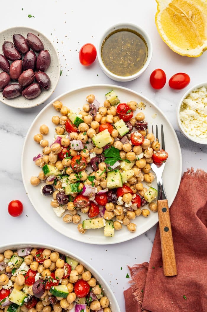 Mediterranean Chickpea Salad served on a white plate with chickpeas, cucumber, cherry tomatoes, red onion, Kalamata olives, feta cheese, and parsley, surrounded by a bowl of dressing, whole cherry tomatoes, a lemon half, crumbled feta, extra olives, a fork with a wooden handle, and a rust-colored linen napkin.