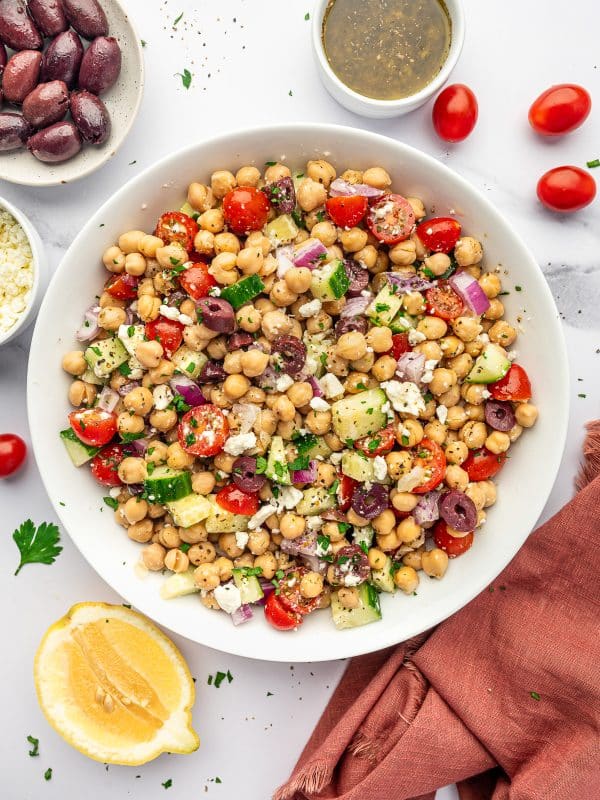 Mediterranean Chickpea Salad in a large white serving bowl with chickpeas, cucumber, cherry tomatoes, red onion, Kalamata olives, crumbled feta cheese, chopped parsley, and lemon oregano dressing, with a halved lemon, extra olives, dressing, tomatoes, and a rust-colored napkin around the bowl.
