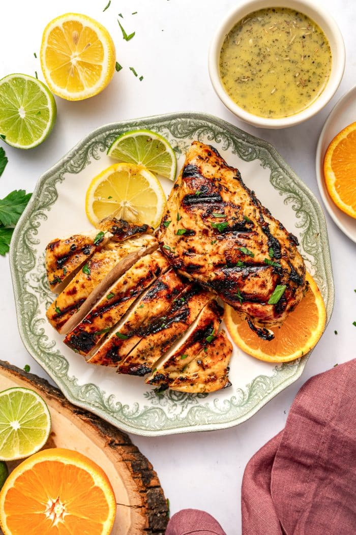 Top-down view of grilled citrus chicken served on a round plate with a pale cream center and decorative sage green border. One chicken breast is sliced into thick pieces, and another whole grilled chicken breast rests beside it. Both pieces have dark grill marks and are garnished with chopped parsley. Lemon, lime, and orange slices are arranged on the plate, and a small off-white bowl of citrus marinade sits above the plate. A halved lemon, halved lime, orange slice, wood slice, parsley, and dusty rose pink linen napkin surround the scene on a white background.