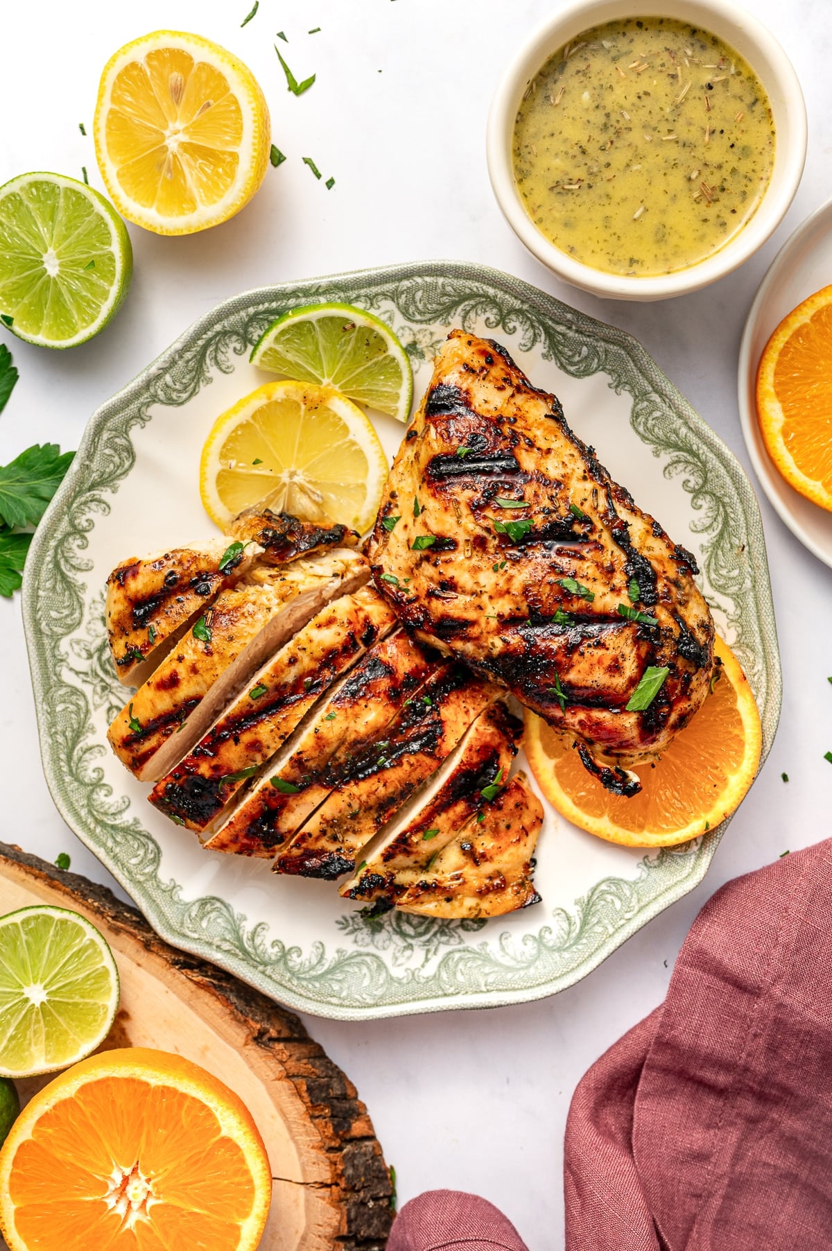 Top-down view of grilled citrus chicken served on a round plate with a pale cream center and decorative sage green border. One chicken breast is sliced into thick pieces, and another whole grilled chicken breast rests beside it. Both pieces have dark grill marks and are garnished with chopped parsley. Lemon, lime, and orange slices are arranged on the plate, and a small off-white bowl of citrus marinade sits above the plate. A halved lemon, halved lime, orange slice, wood slice, parsley, and dusty rose pink linen napkin surround the scene on a white background.
