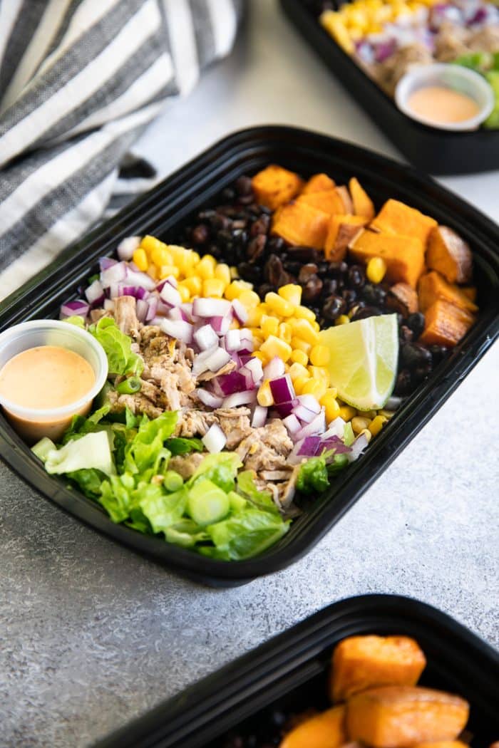 Side angle of a plastic meal prep tray filled with green chili verde pork, onion, beans, roasted sweet potatoes, and lettuce.