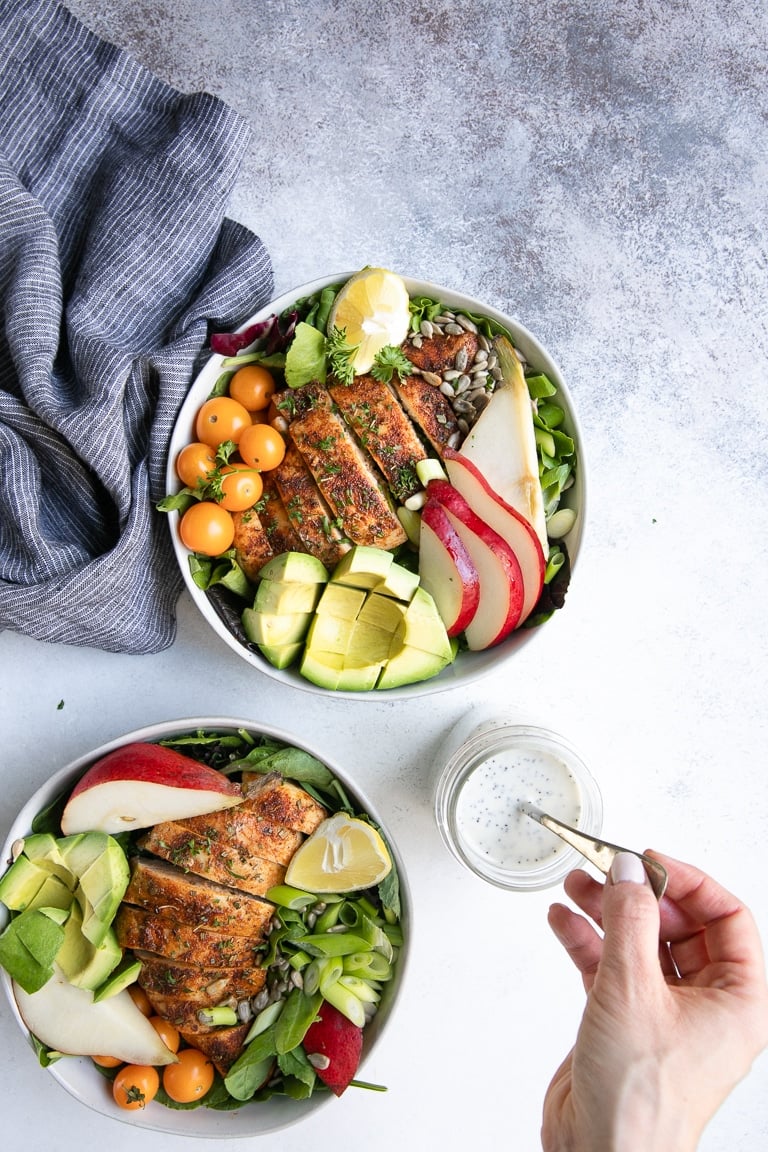 Two white bowls filled with salad greens, cooked chicken breast, pears, and avocado ready to be drizzled with poppyseed dressing.