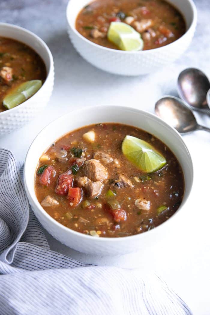 Several white bowls filled with easy pork green chili stew made with mild green chiles, tomatoes, pork shoulder, and garnished with a lime wedge.