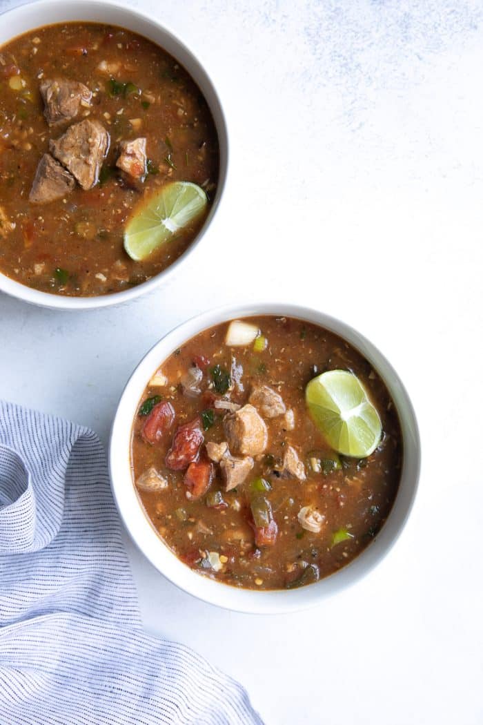 Overhead image of two white bowls filled with easy pork green chili stew made with mild green chiles, tomatoes, chunks of tender pork shoulder, and garnished with a lime wedge.