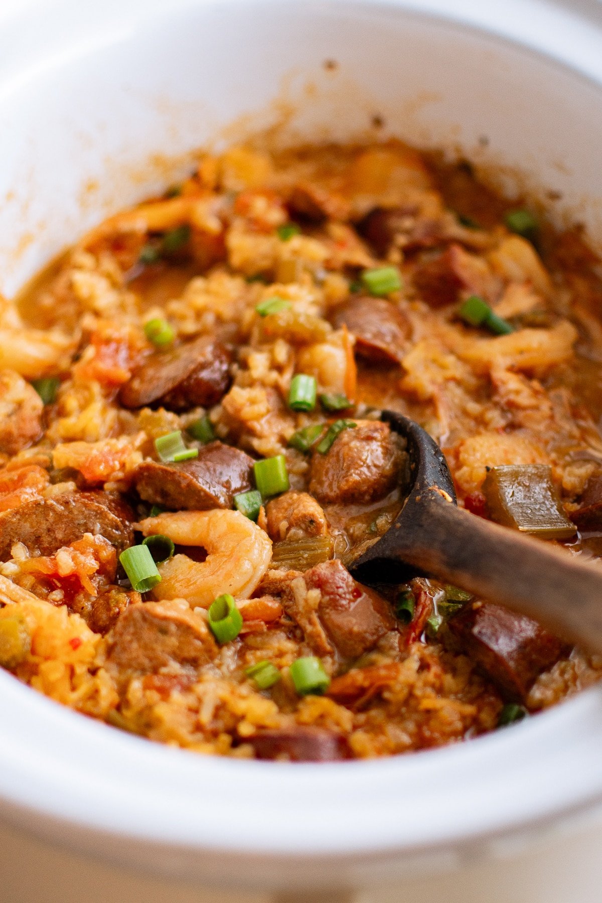 Finished jambalaya in a white slow cooker garnished with green onions and served with a wooden spoon.