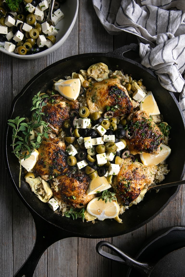 Large cast iron skillet filled with cooked bone-in skin-on chicken thighs set atop lemon rice and garnished with sliced olives and feta cheese.