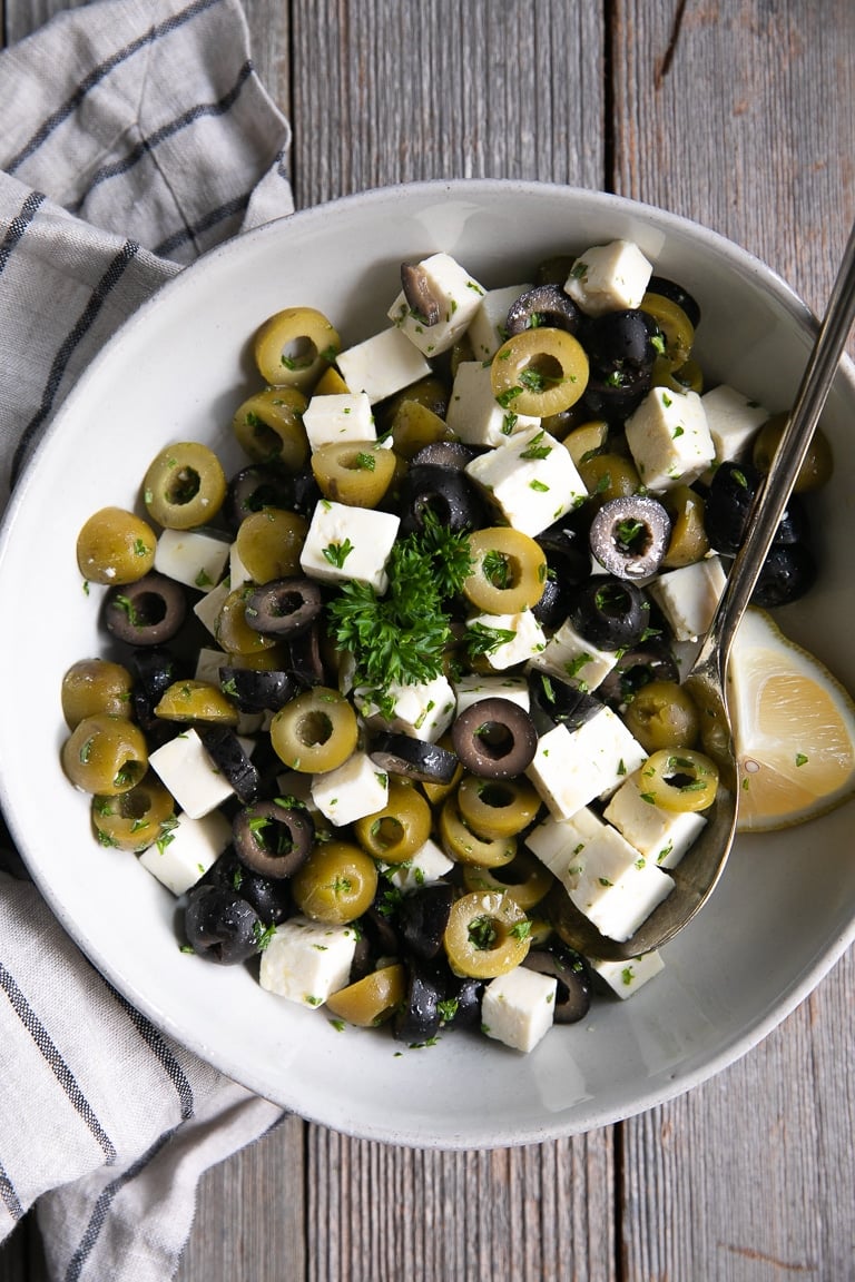 Large white bowl filled with cubes of feta cheese, sliced olives, fresh herbs, and lemon juice.
