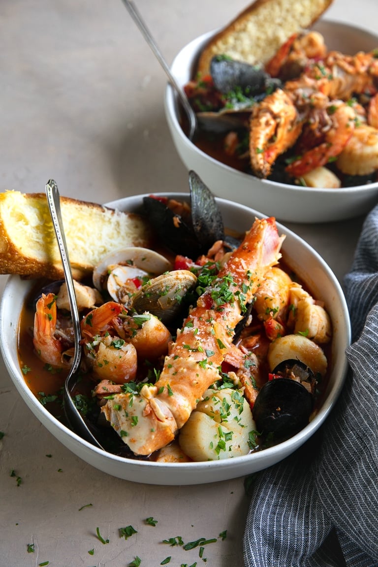 Two bowls of Cioppino seafood stew filled with shrimp, scallops, crab legs, fish, clams, and mussels and served with slices of bread.