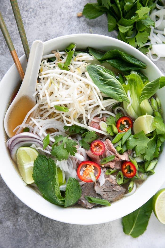 Tazón blanco lleno de caldo de pho, brotes de judías mungo, menta fresca, albahaca tailandesa, cebollas y carne de vacuno.