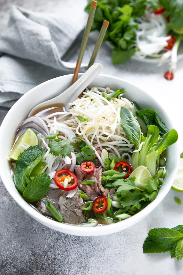 White bowl filled with homemade pho garnished with herbs, fresh greens, and filled with rice noodles.