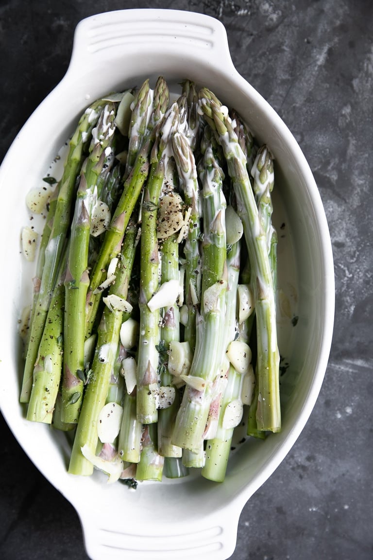 Raw asparagus,garlic, and cream in a white oval baking dish.