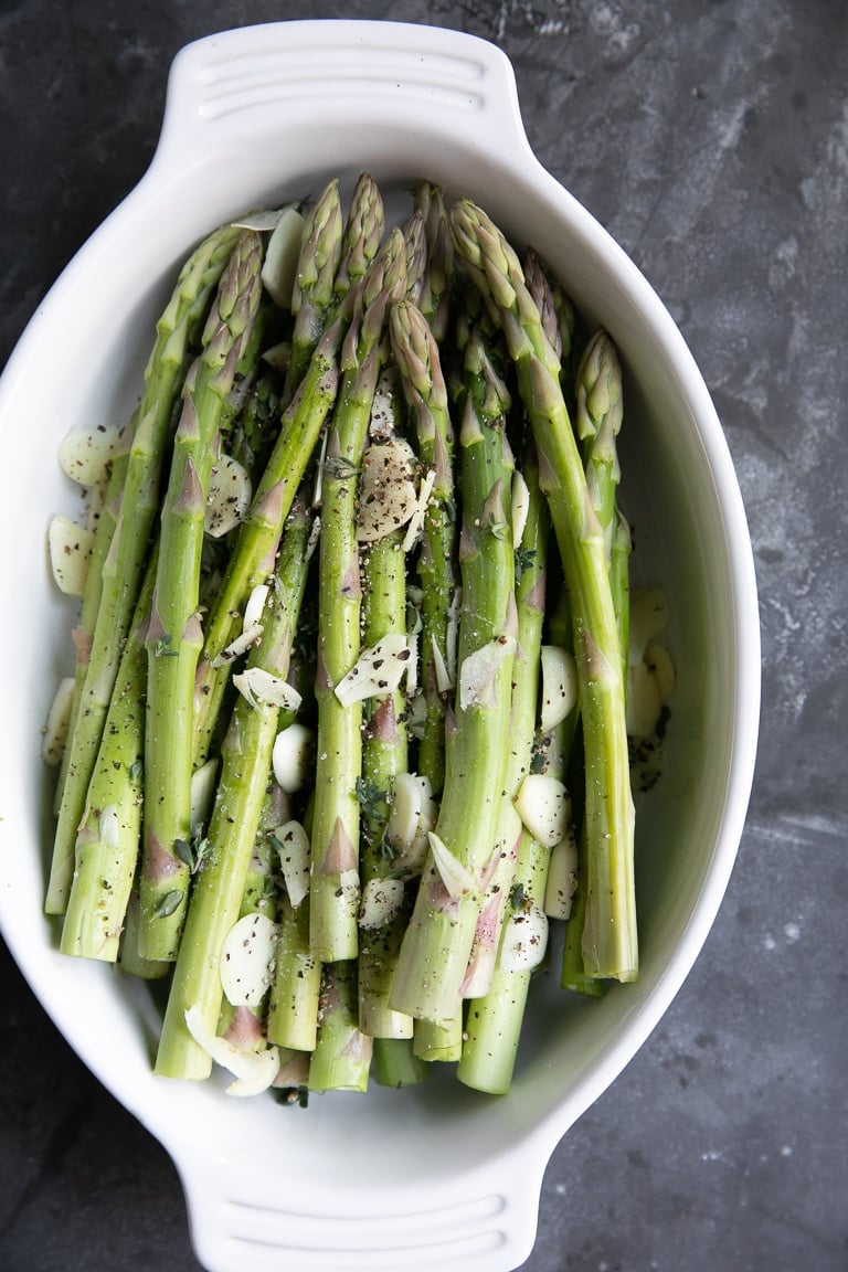 Raw asparagus and garlic in a white oval baking dish.