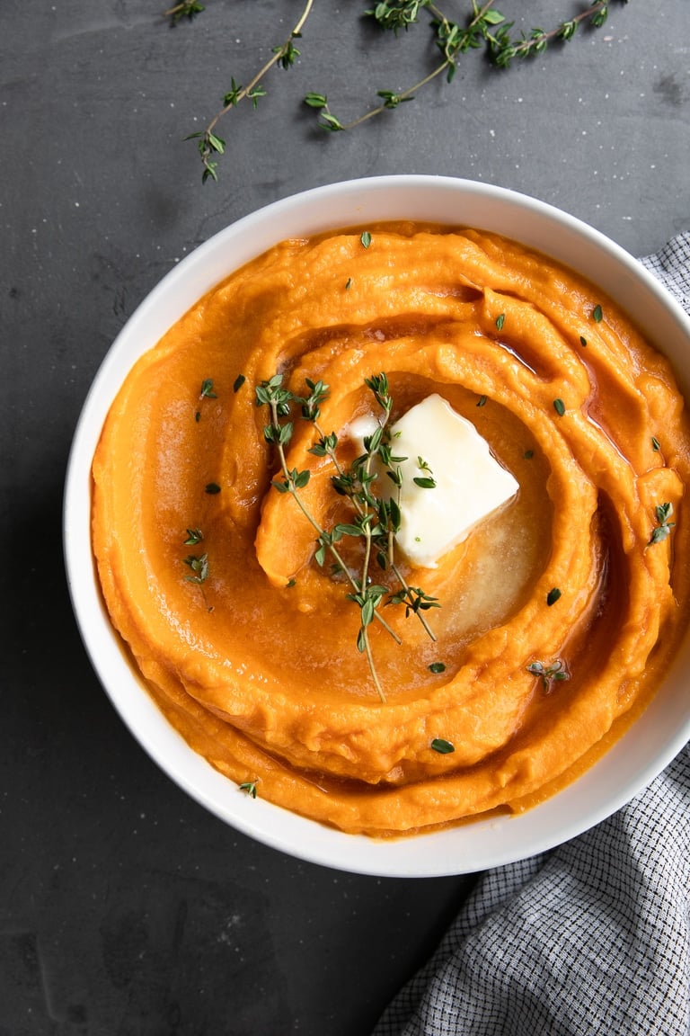 White bowl filled with creamy mashed sweet potatoes topped with butter and fresh thyme