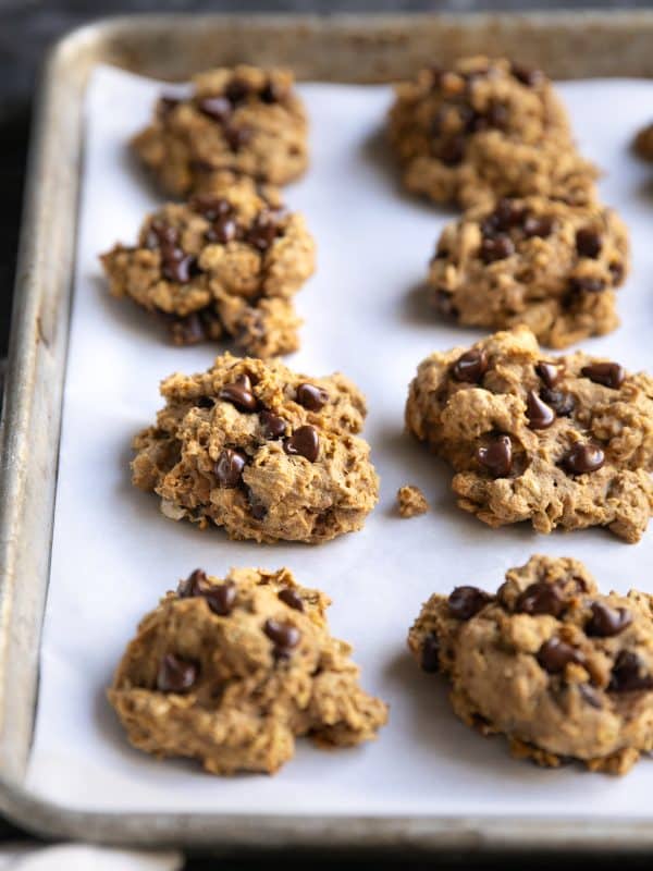 Freshly baked pumpkin oatmeal chocolate chip cookies on a white parchment-lined metal baking sheet, photographed at an angle with several thick cookies in focus and melted chocolate chips scattered across their tops.