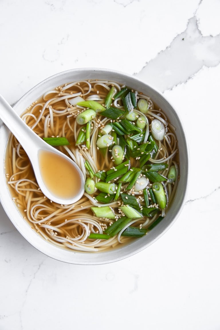 White bowl filled with bone broth, cooked soba noodles, and chopped green onions.