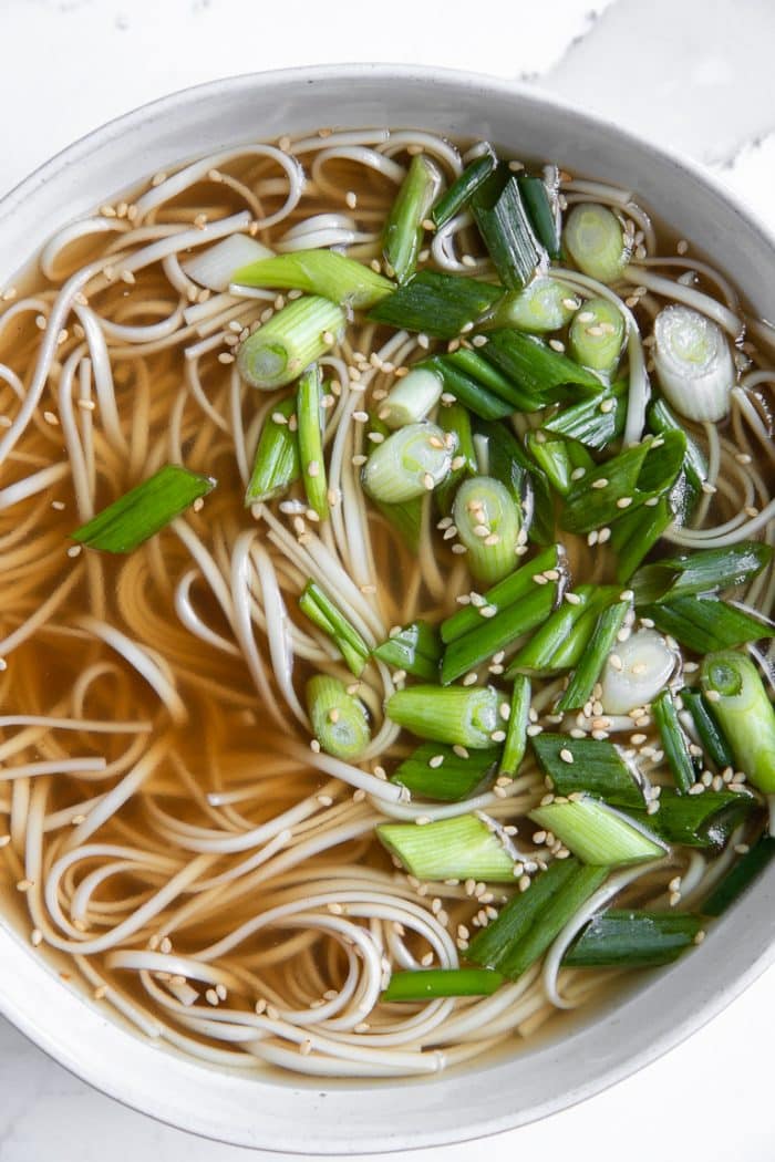 White bowl filled with bone broth, cooked soba noodles, and chopped green onions.
