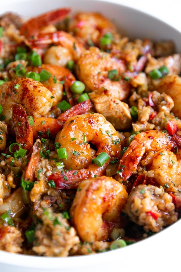 Close up image of a large white pot filled with cooked jambalaya made with white rice, chicken, andouille sausage, and shrimp, and garnished with chopped parsley and green onions.