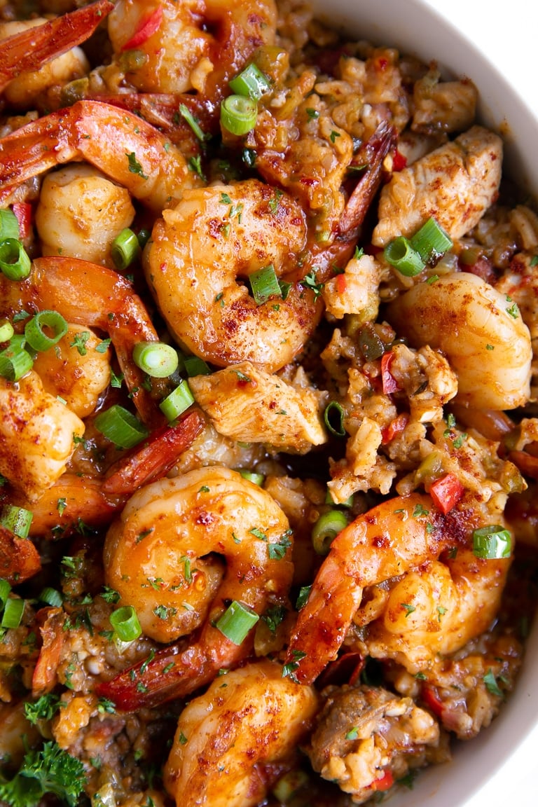 Close up image of a large white pot filled with cooked jambalaya made with white rice, chicken, andouille sausage, and shrimp, and garnished with chopped parsley and green onions.