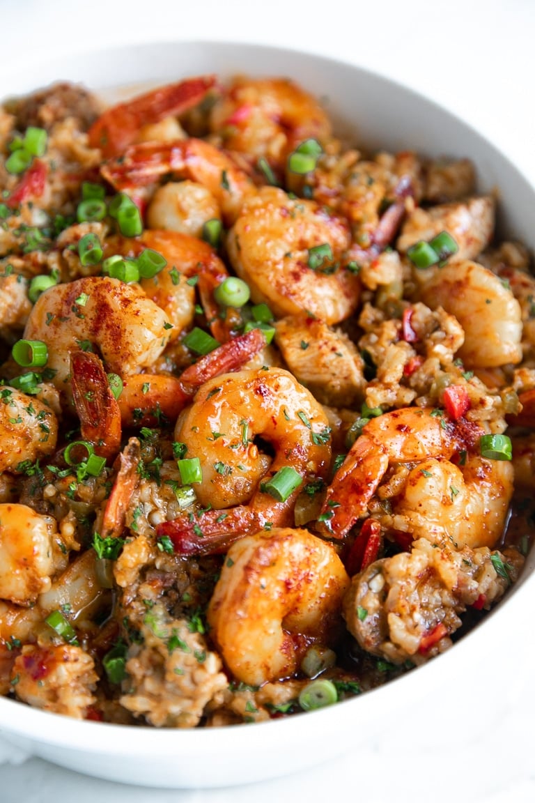 Large white pot filled with cooked jambalaya made with white rice, chicken, andouille sausage, and shrimp, and garnished with chopped parsley and green onions.
