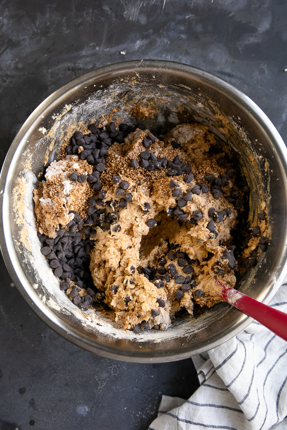 Thick pumpkin oatmeal cookie dough in a stainless steel mixing bowl topped with semi-sweet chocolate chips and whole toasted flaxseeds, with a red rubber spatula resting along the side.