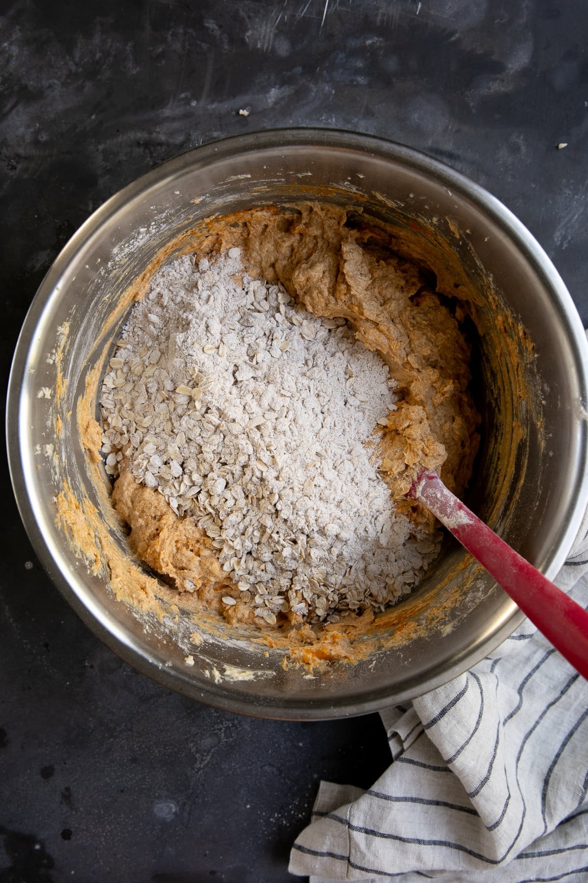 Pumpkin oatmeal chocolate chip cookie dough in a large stainless steel mixing bowl with flour and rolled oats added on top of the orange-brown pumpkin mixture and a red rubber spatula resting inside.