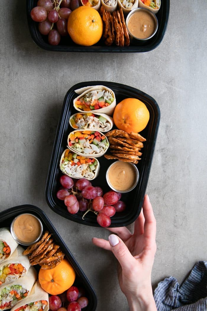 Thai chicken wrap meal prep trays filled with grapes, pretzels, and oranges.