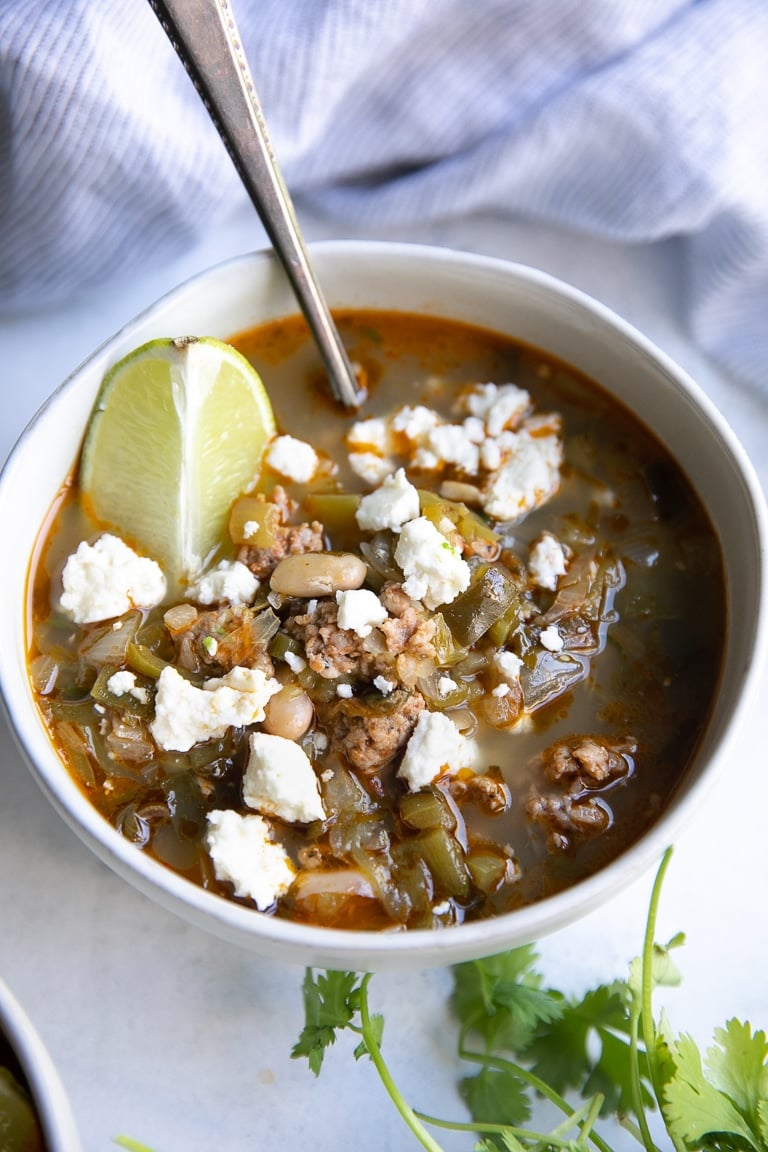 Close-up image of a white bowl filled with pork green chile made with ground pork, green chiles, onion, and garnished with lime and cotija cheese.