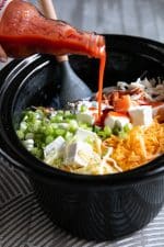 Ingredients for Buffalo Chicken dip being put into the slow cooker, with the hot sauce being poured