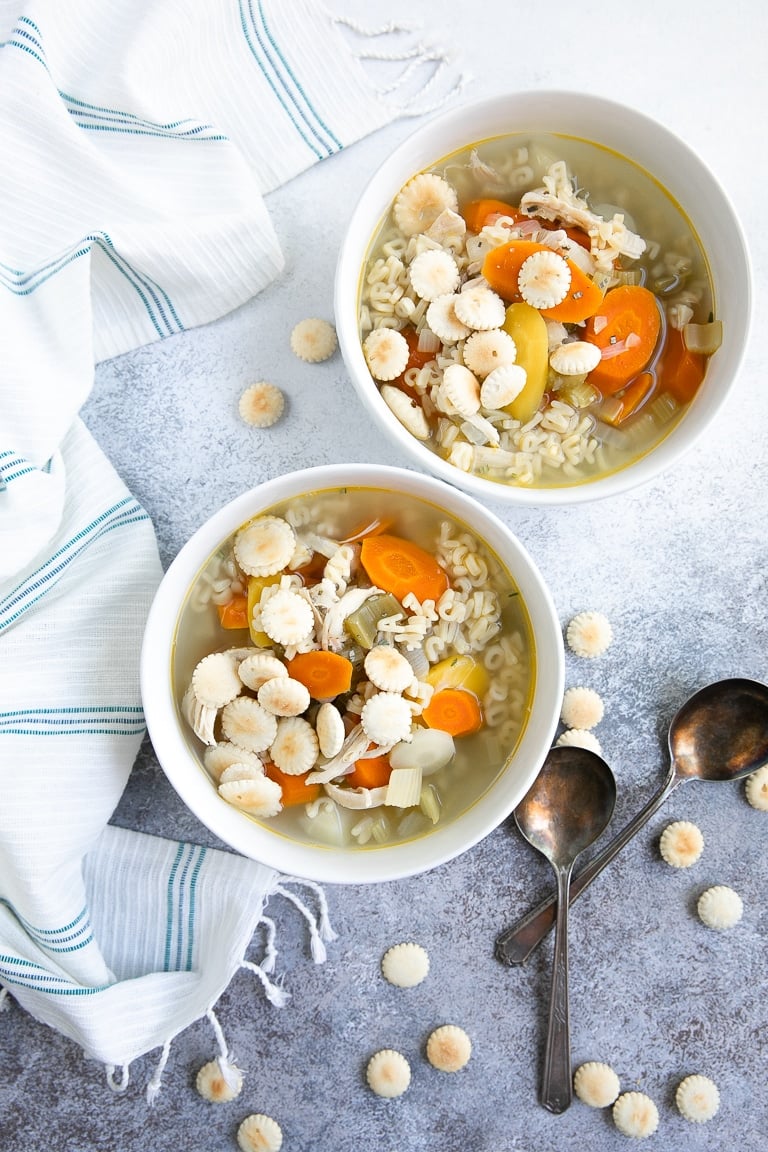 Two white bowls of alphabet chicken soup topped with salty oyster crackers next to a white dish cloth.