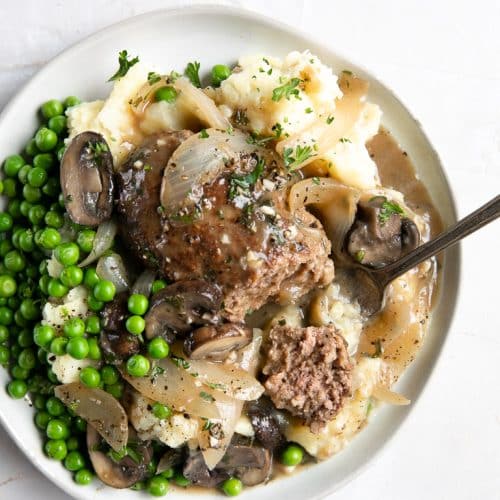 White plate filled with Salisbury steak beef patty on top of creamy mashed potatoes topped with onion and mushroom gravy served with a side of buttered peas.