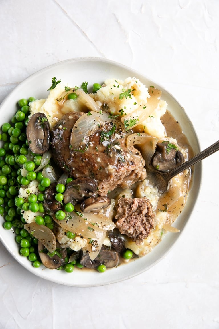 White plate filled with Salisbury steak beef patty on top of creamy mashed potatoes topped with onion and mushroom gravy served with a side of buttered peas.