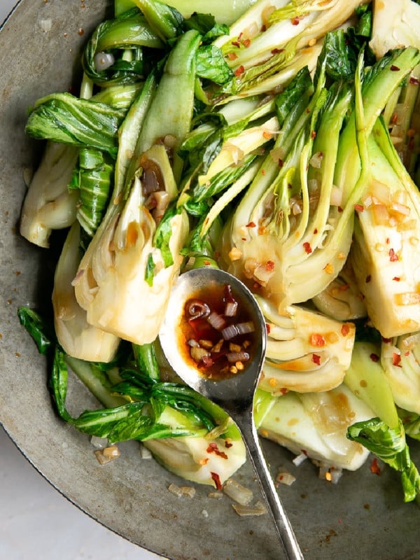 Stir-fried baby bok choy with garlic, shallots, and red pepper flakes in a skillet with a spoonful of savory soy-based sauce.