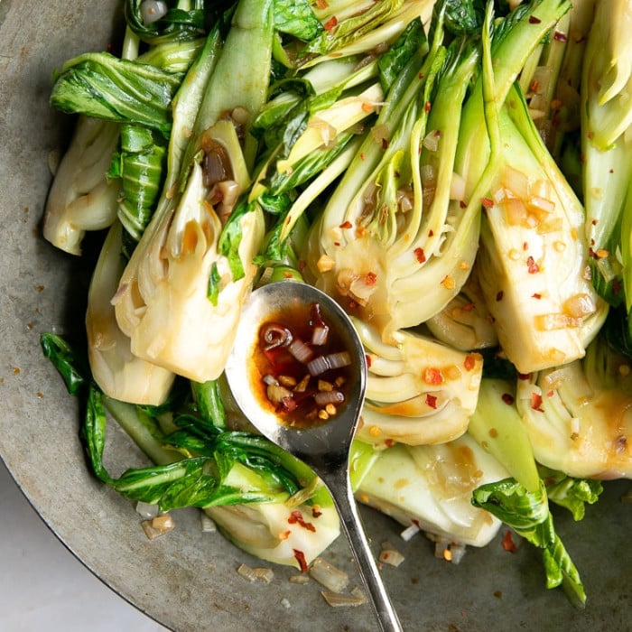Stir-fried baby bok choy with garlic, shallots, and red pepper flakes in a skillet with a spoonful of savory soy-based sauce.