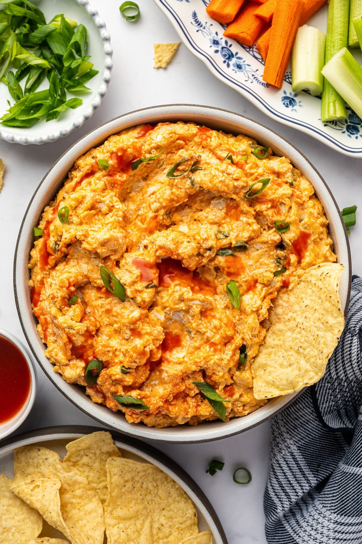 Creamy Buffalo Chicken Dip in a serving bowl topped with chopped green onions and hot sauce, surrounded by tortilla chips, carrot sticks, celery sticks, and additional sliced green onions.