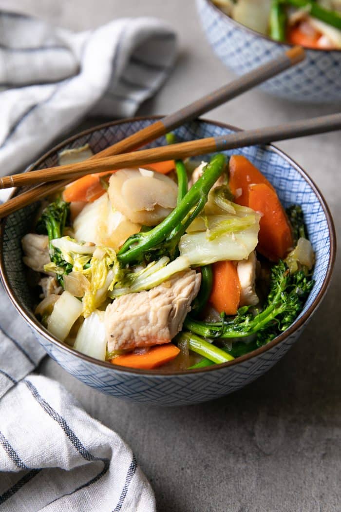 Blue and white bowl filled with chicken, cabbage, broccoli stir fry with chopsticks resting on top.