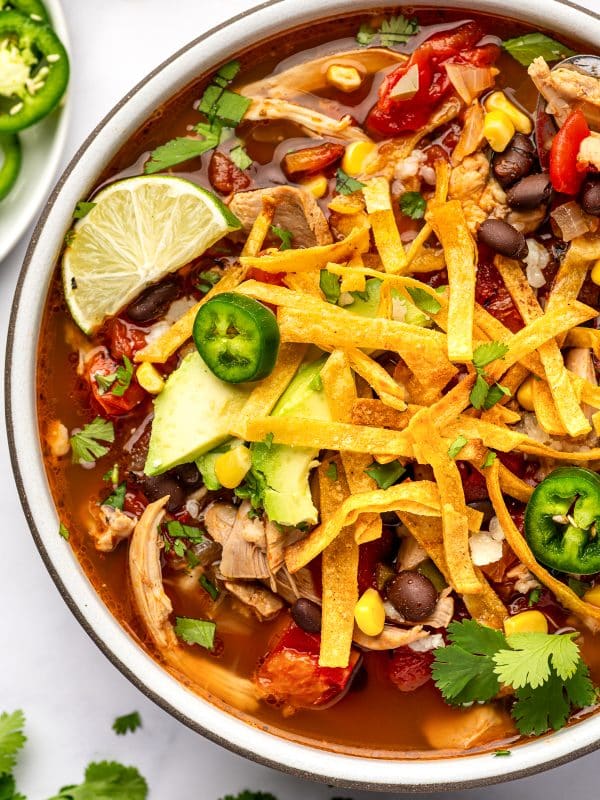 A white bowl filled with chunky chicken tortilla soup in a reddish-orange broth. Golden baked tortilla strips are piled across slices of green avocado, with fresh jalape&ntilde;o rings, chopped cilantro, and a lime wedge among the toppings. Shredded chicken, black beans, yellow corn, and pieces of red tomato are visible beneath and around the crispy strips.
