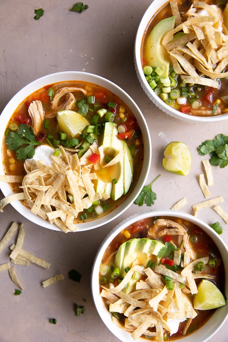 Three bowls filled with chicken tortilla soup and garnished with tortilla strips, avocado, lime, and sour cream.