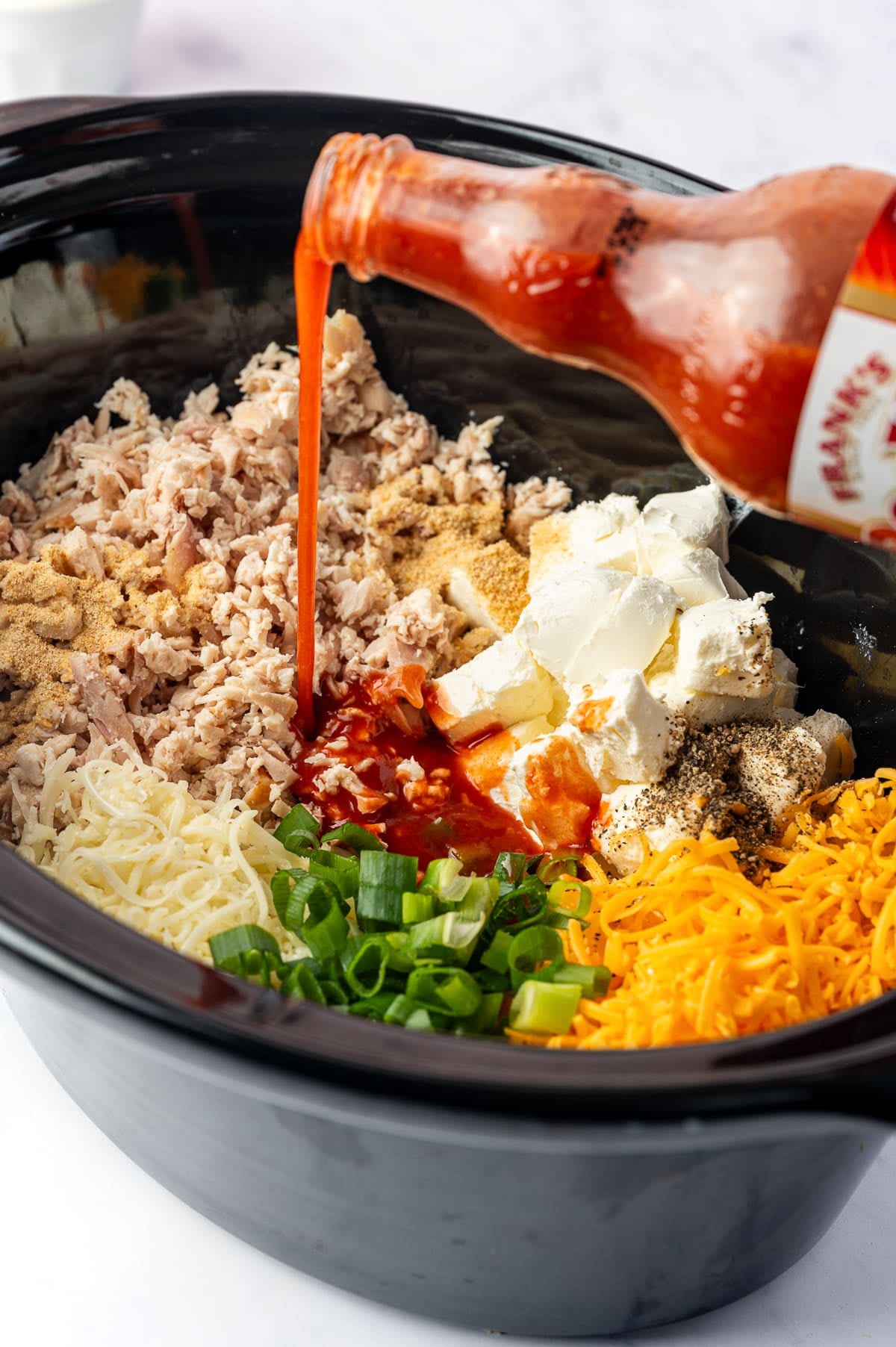 Frank's RedHot Original being poured into a slow cooker filled with shredded chicken, cubed cream cheese, shredded cheddar and mozzarella, chopped green onions, and seasonings for Crock Pot Buffalo Chicken Dip.