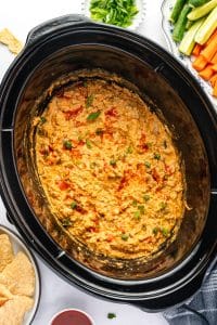 Finished Crock Pot Buffalo Chicken Dip topped with chopped green onions and drizzled with hot sauce, served directly from the slow cooker with tortilla chips, carrot sticks, celery sticks, and extra green onions nearby.