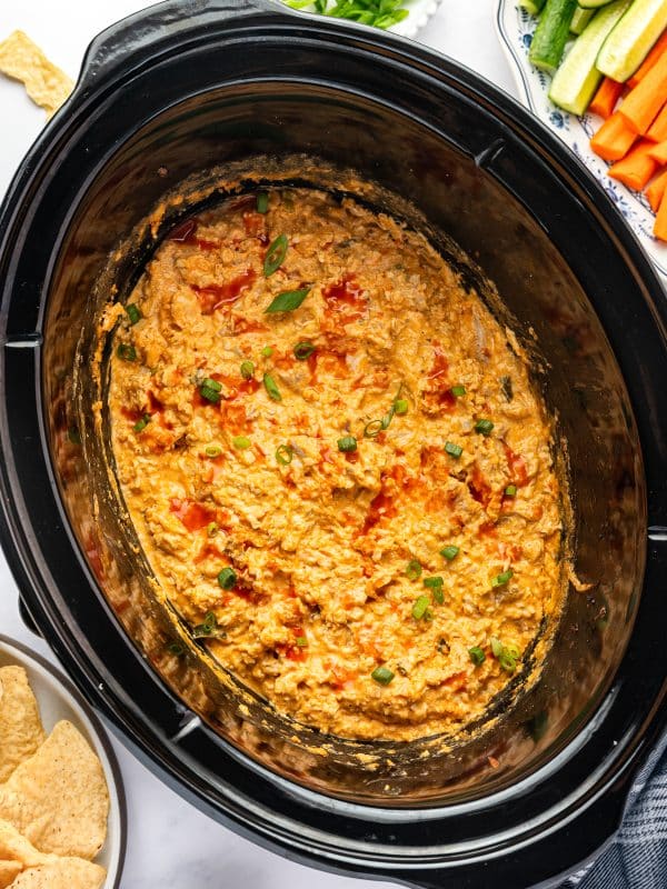 Finished Crock Pot Buffalo Chicken Dip topped with chopped green onions and drizzled with hot sauce, served directly from the slow cooker with tortilla chips, carrot sticks, celery sticks, and extra green onions nearby.