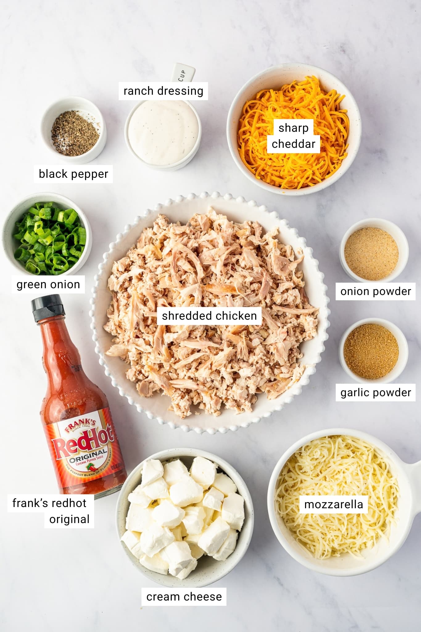 Ingredients for Crock Pot Buffalo Chicken Dip labeled individually, including shredded chicken, cubed cream cheese, shredded mozzarella, sharp cheddar, Frank's RedHot Original, ranch dressing, green onions, garlic powder, onion powder, and black pepper.