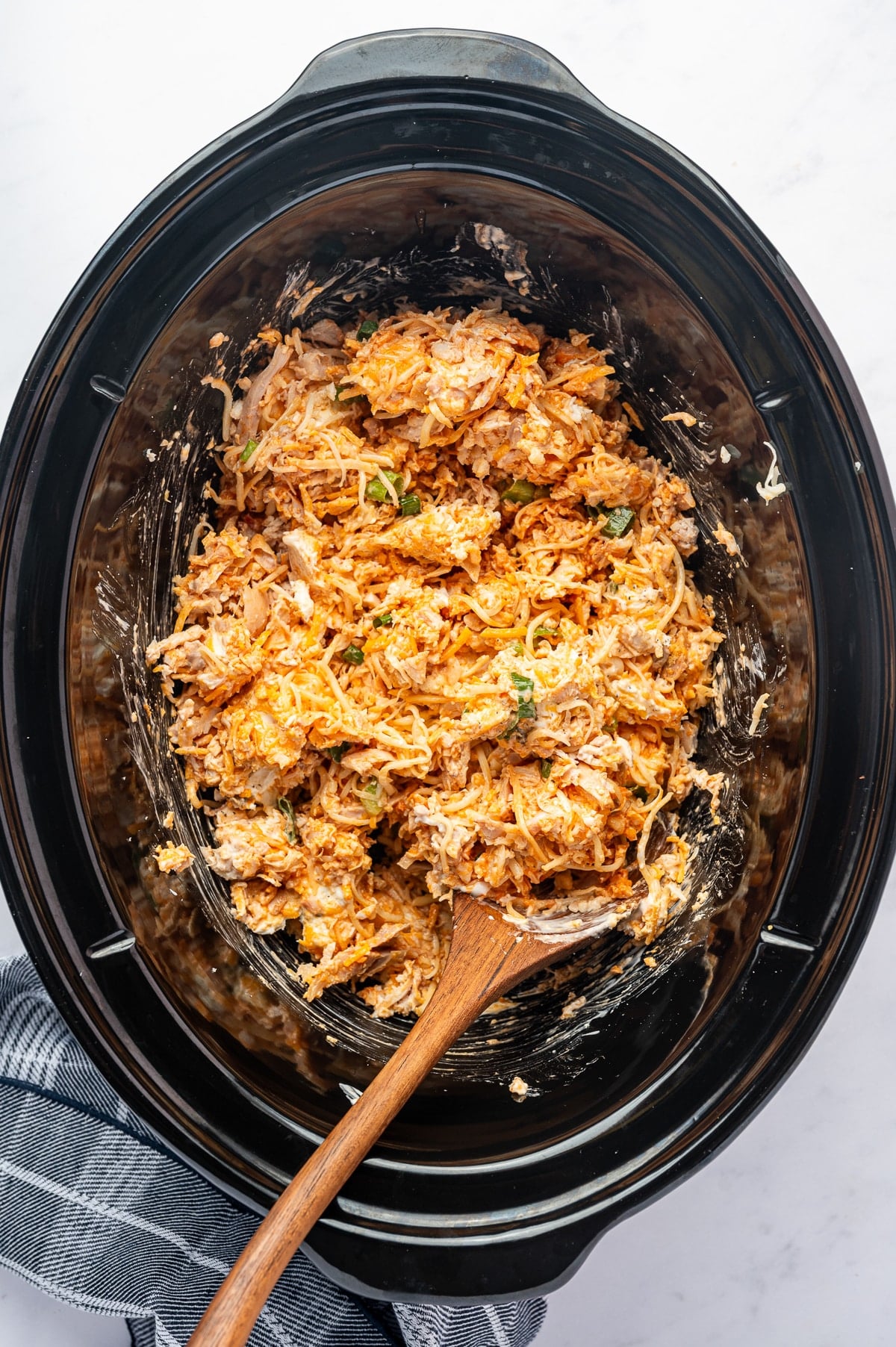 Shredded chicken, cream cheese, cheddar, mozzarella, green onions, hot sauce, and seasonings partially mixed together in a slow cooker before cooking, with a wooden spoon resting in the mixture.