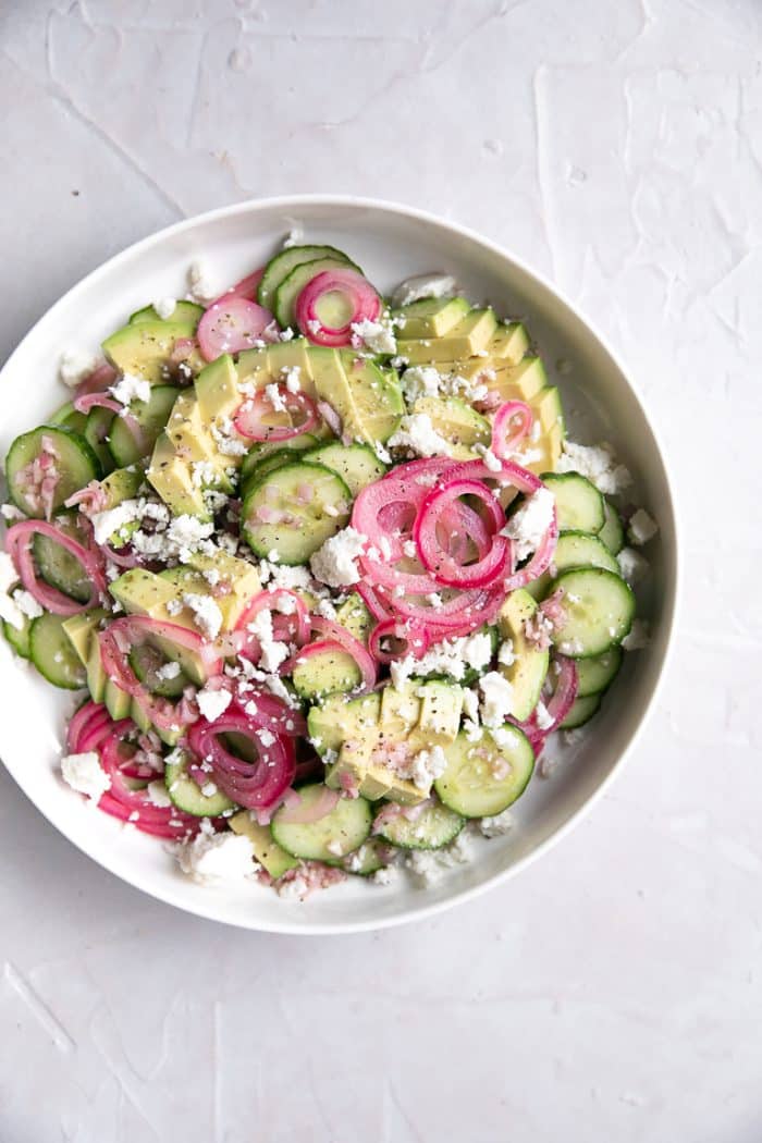 Large white salad plate filled with sliced cucumbers, avocado, and pickled onions covered in crumbled casero cheese and homemade vinaigrette.